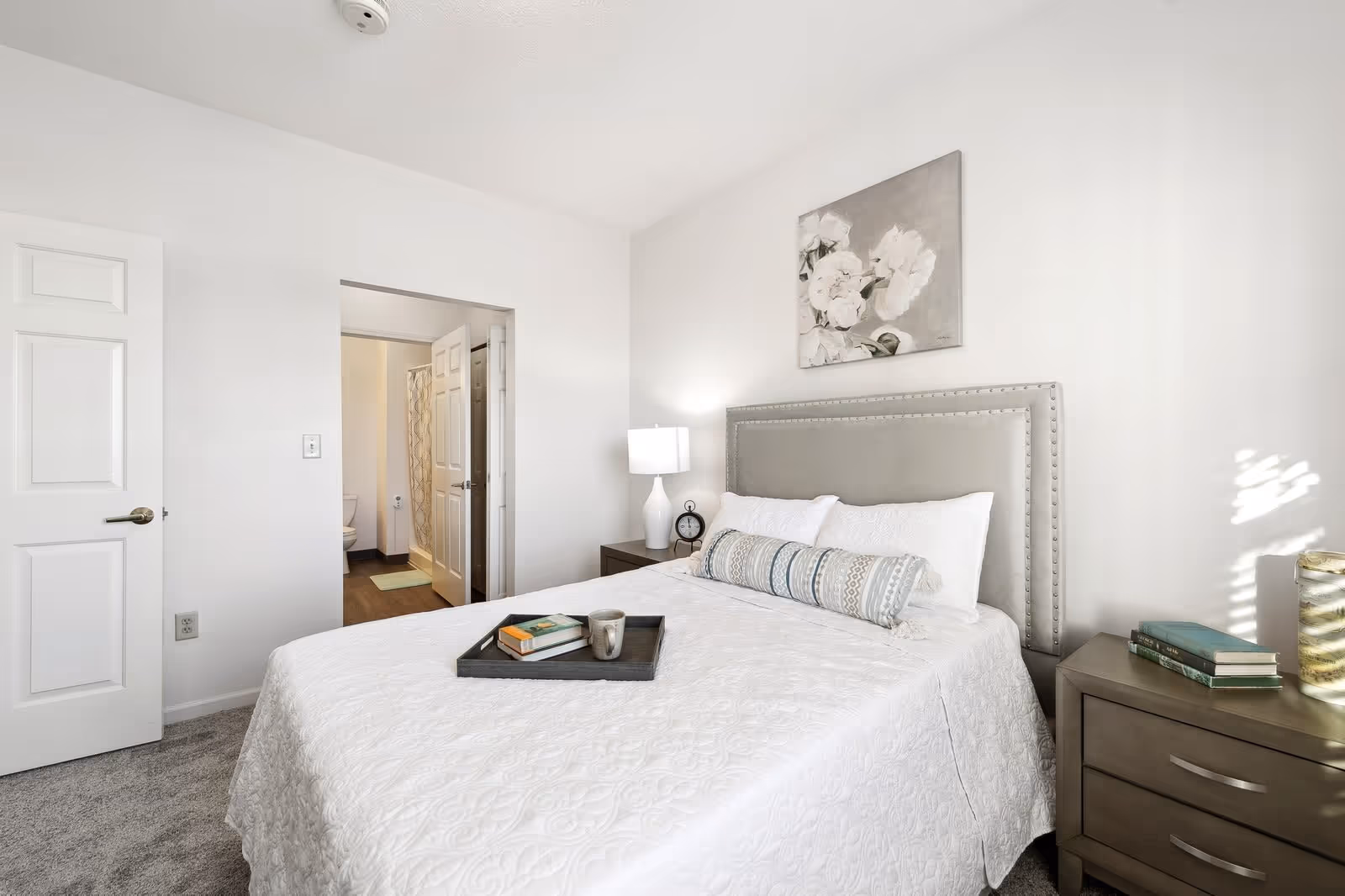 A bright bedroom with a neatly made bed featuring a white quilt and decorative pillows. A tray with books and a mug rests on the bed. There is a nightstand with a lamp and an alarm clock on one side, and another nightstand with books and a decorative vase on the other. A doorway leads to a bathroom with a visible toilet and shower curtain. The walls are white, and a floral painting hangs above the bed.