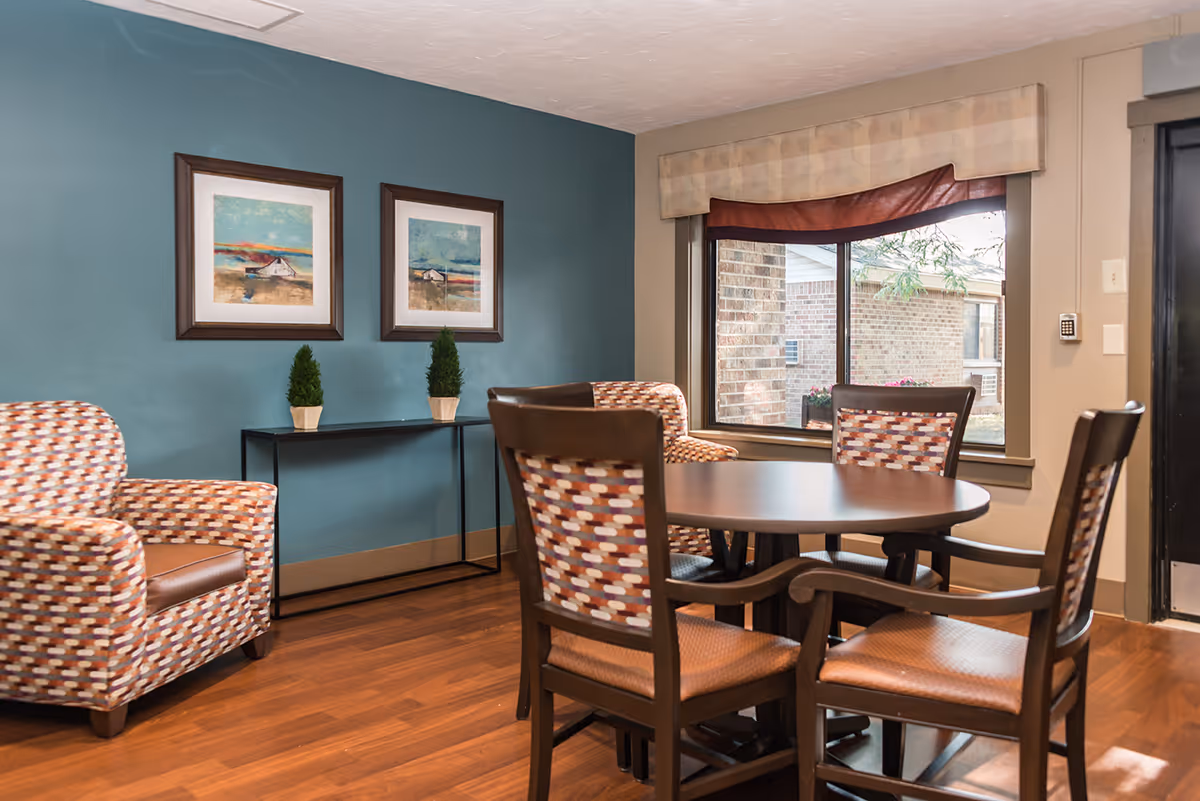 A cozy seating area in a senior living facility featuring a round wooden table surrounded by four chairs with patterned upholstery. Two matching armchairs are placed against a teal accent wall, which is decorated with two framed landscape paintings and a narrow black console table holding two small potted plants. A large window with a beige and brown valance lets in natural light and shows an exterior brick wall outside.