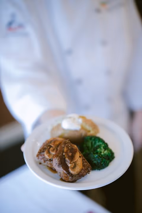 A chef in a white uniform holding a white plate with a meal consisting of two pieces of steak topped with mushrooms, a baked potato with sour cream, and a serving of steamed broccoli.