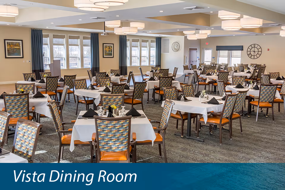 A spacious dining room with multiple tables covered in white tablecloths, each set with black napkins, glasses, and small flower arrangements. The room has patterned chairs with orange seats, large windows with blue curtains, and modern ceiling lights. The walls are decorated with framed pictures and a large clock.