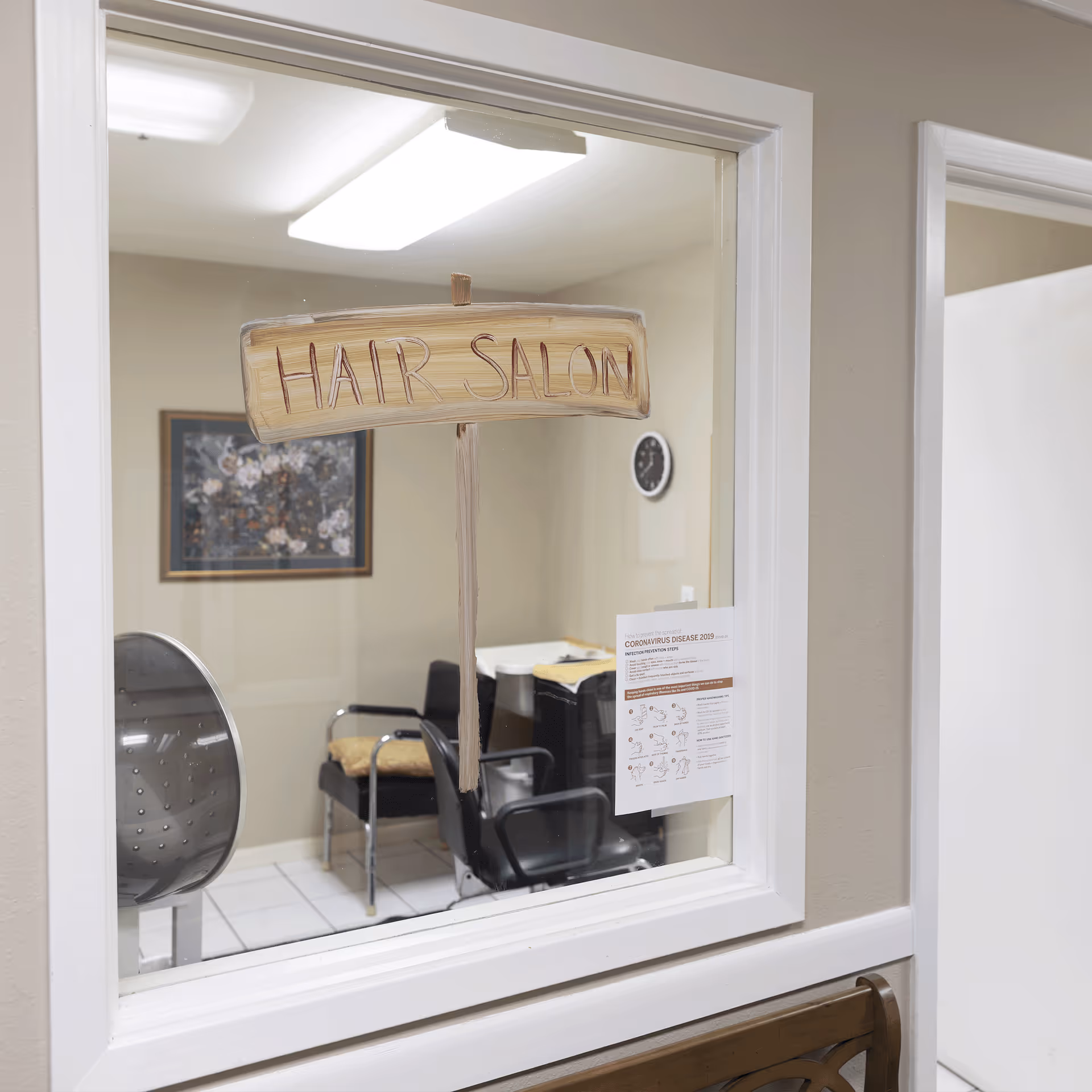 View through a window into a hair salon room with chairs, a hair dryer, a wall clock, and a framed floral picture. A wooden sign reading 'HAIR SALON' is attached to the window. A notice about Coronavirus Disease 2019 is posted on the window.