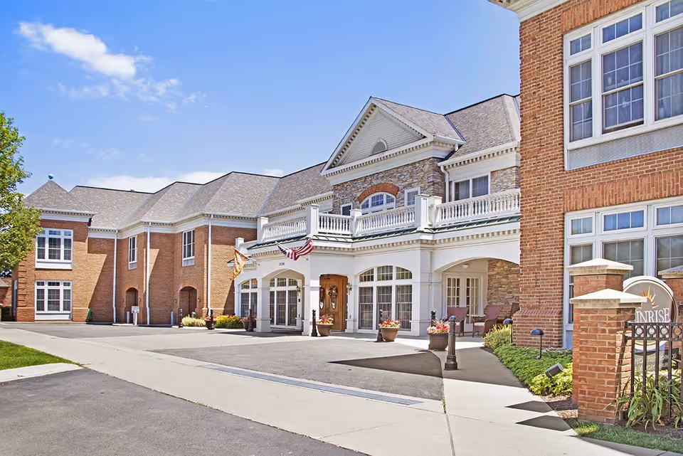 Front exterior of a red-brick senior living building with a covered entrance, balcony, flags, and potted plants.