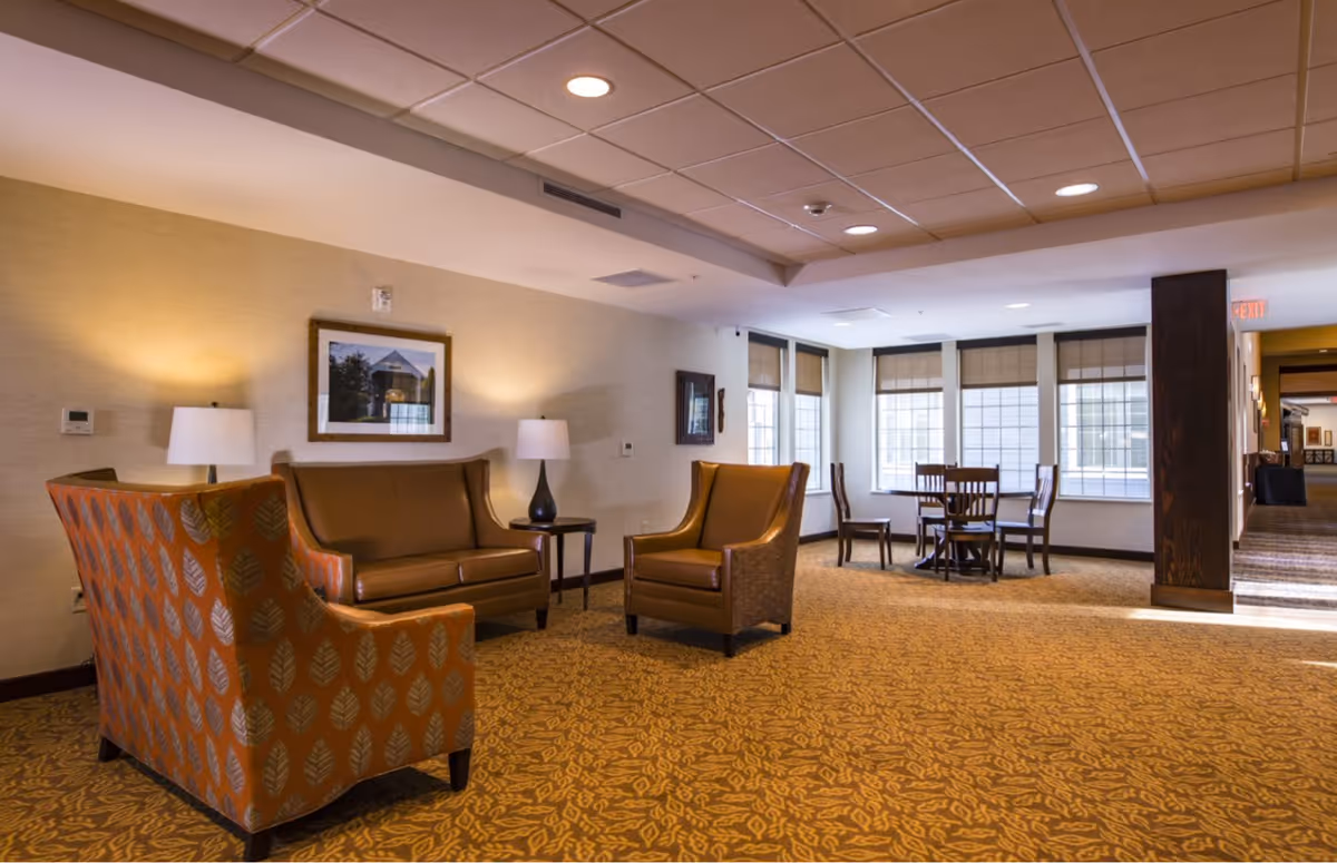 Well-lit senior living lounge with upholstered chairs, a sofa, side tables and lamps, and a small dining table by windows.