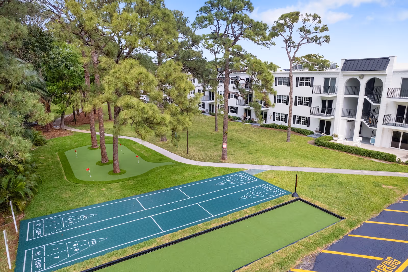 Outdoor recreational area at Independence Hall Senior Living featuring shuffleboard courts, a putting green with several holes, surrounded by tall pine trees and adjacent to a white multi-story residential building with balconies and staircases.