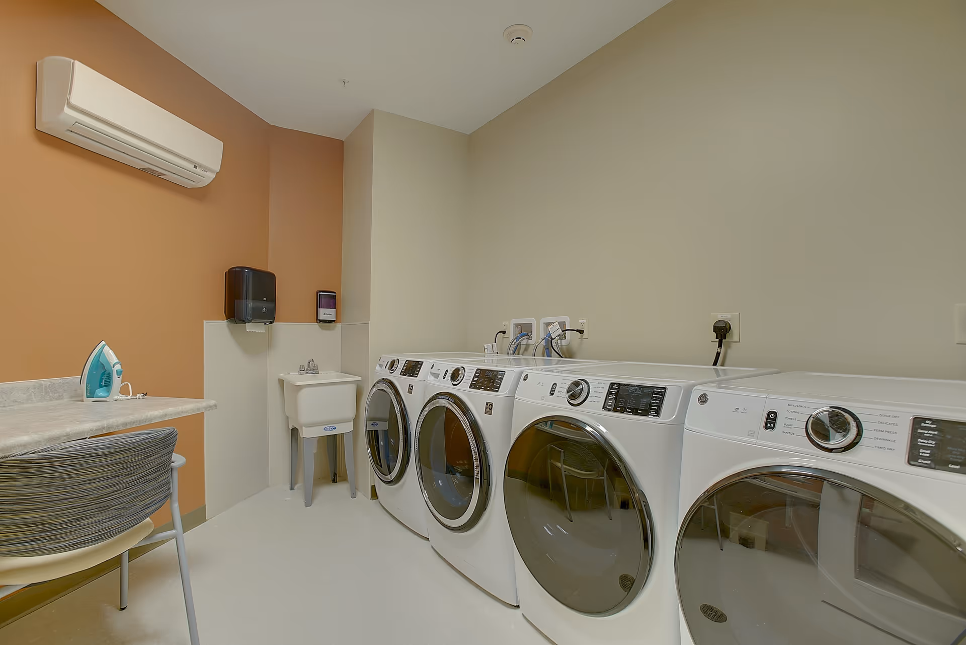 Laundry room with four front-loading washing machines and dryers lined up against a beige wall. There is a small utility sink in the corner, a countertop with an iron on it, and a chair with a striped backrest. The walls are painted in beige and light brown tones, and there is an air conditioning unit mounted on the wall.