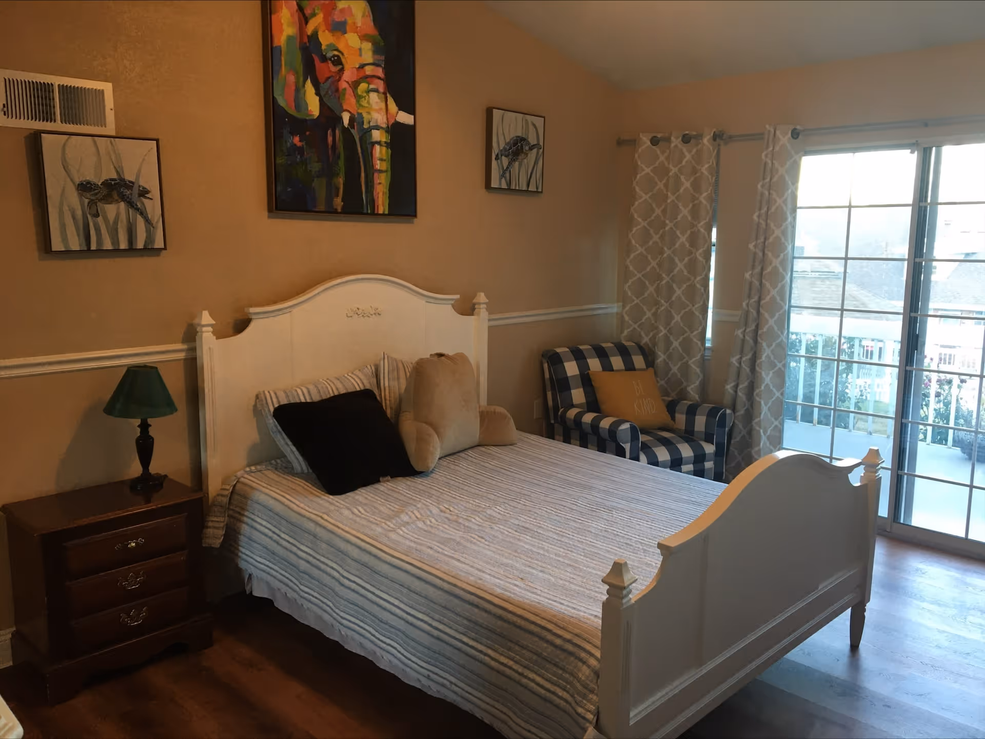A bedroom with a white wooden bed frame, striped bedding, and several pillows including a teddy bear-shaped pillow. There is a wooden nightstand with a green lamp on the left side of the bed. On the right side, there is a blue and white checkered armchair with a yellow pillow. The room has beige walls with three framed pictures, and a large sliding glass door with patterned curtains letting in natural light.