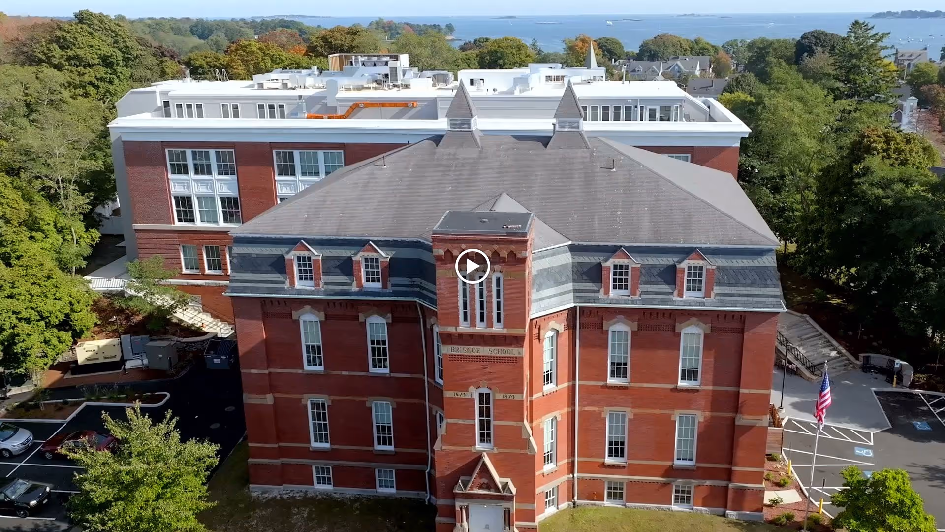 Aerial view of a large red brick building with multiple windows and a gray roof, surrounded by trees and parking areas. The building has a sign that reads 'Briscoe School' and is part of a larger complex with a modern white building behind it. An American flag is visible near the parking lot, and the background shows a distant view of water and more greenery.