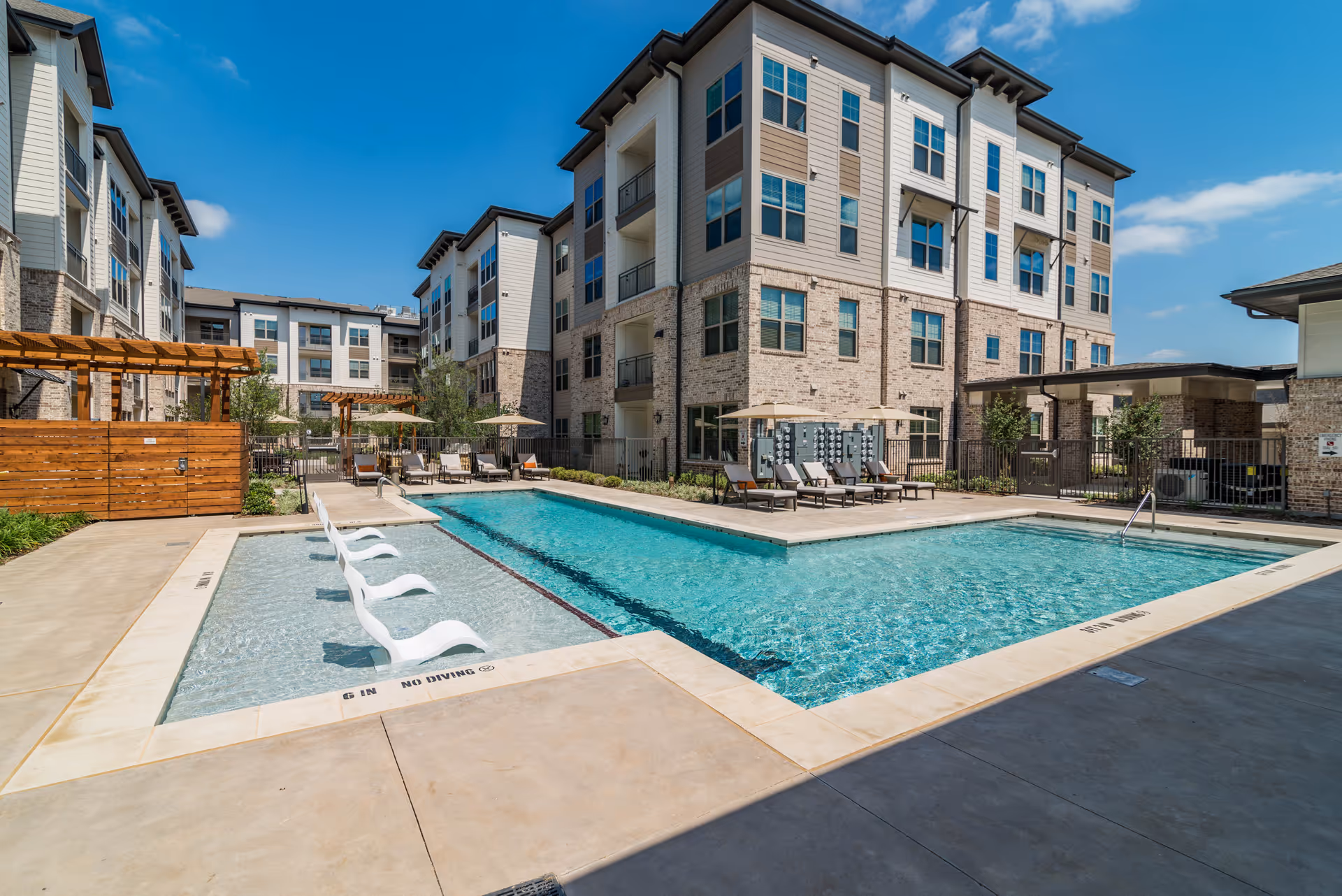 Outdoor swimming pool area at Album Keller Ranch with lounge chairs and umbrellas around the pool, surrounded by multi-story residential buildings under a clear blue sky.
