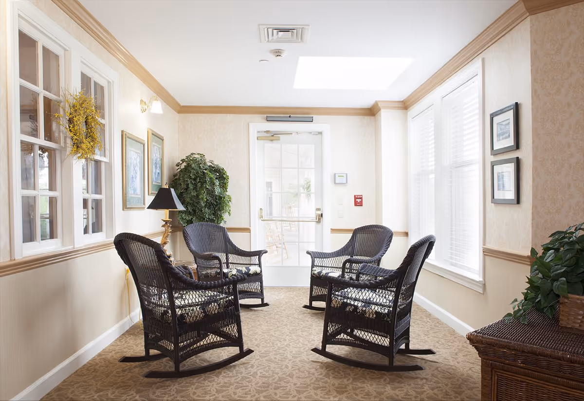 Sunlit common sitting area with four black wicker rocking chairs, potted plants, framed art, and a glass door.