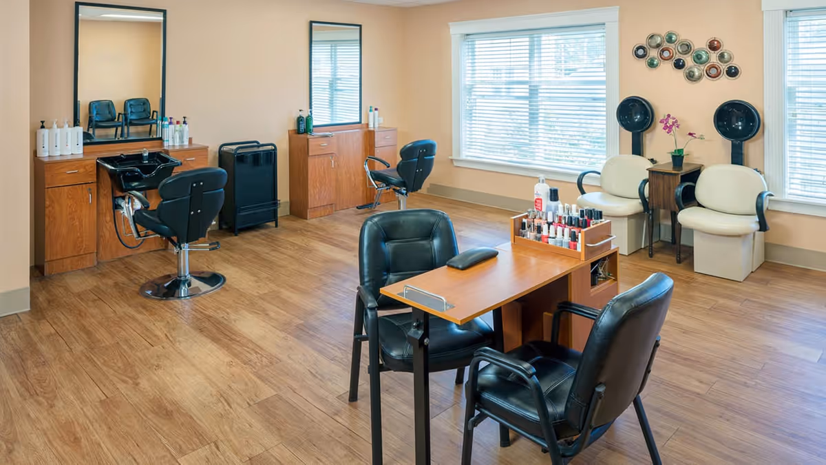 A well-lit salon room with wooden flooring featuring two black salon chairs in front of mirrors and sinks, a small table with nail polish bottles and two black chairs, and two white chairs with hair drying hoods near the windows.
