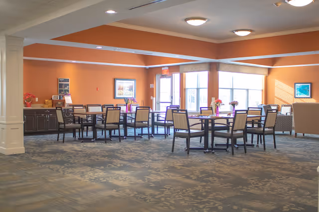 Spacious communal dining area with multiple tables and chairs, orange walls, and large windows letting in natural light.