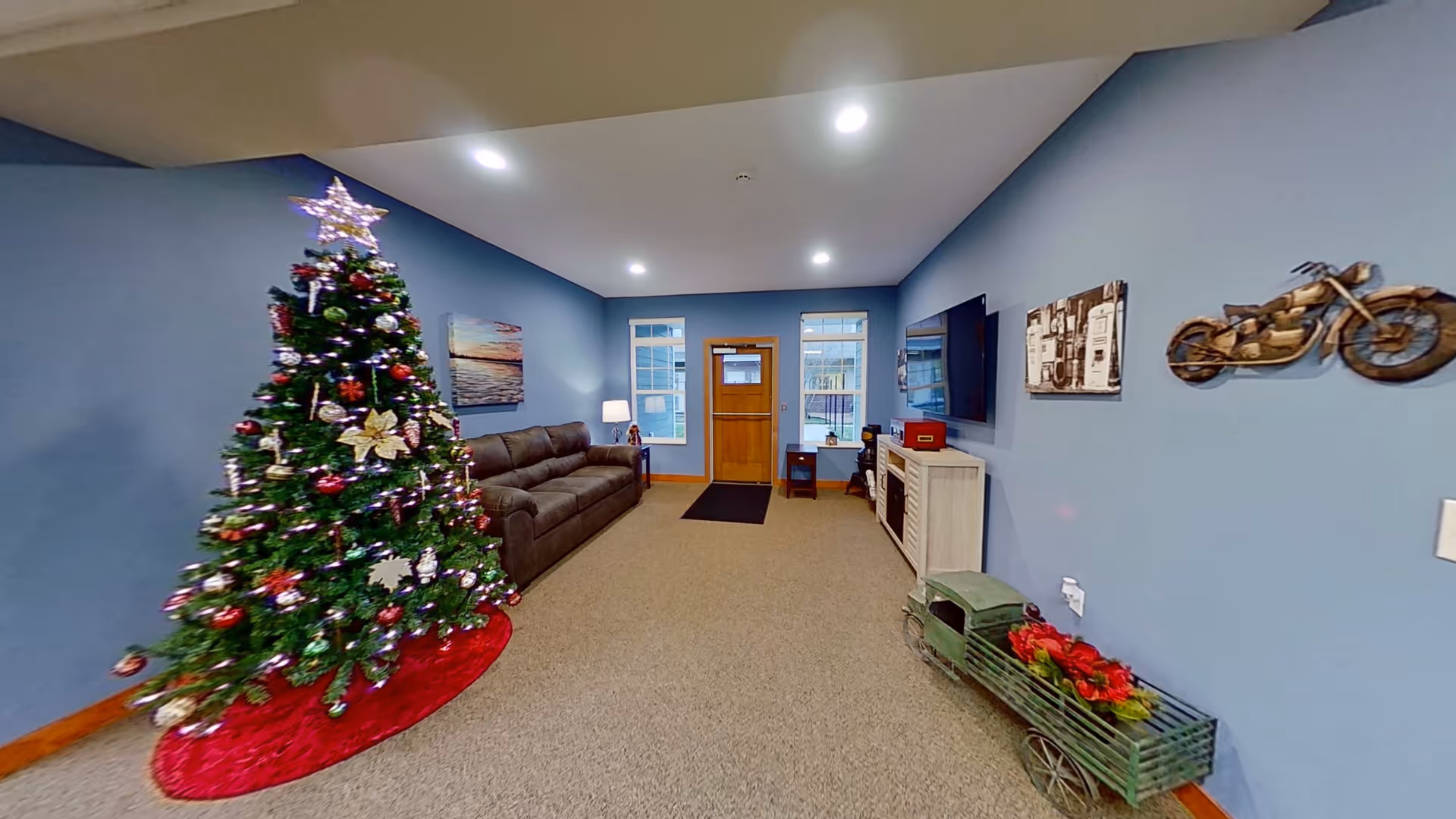 A cozy living room with blue walls featuring a decorated Christmas tree with lights and ornaments on a red tree skirt. There is a brown sofa against the left wall, a wooden door with two windows on either side at the far end, and a flat-screen TV mounted on the right wall above a white cabinet. The room is decorated with wall art including a motorcycle sculpture and a vintage gas pump image. A small green decorative cart with red flowers is on the floor near the right wall.