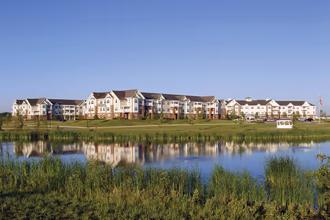 Wide exterior view of a large multi-story senior living facility named American House Cedarlake, with a pond and grassy area in the foreground reflecting the building under a clear blue sky.