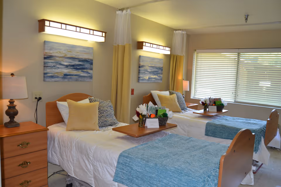 A bright shared patient room with two made hospital beds, bedside tables, overbed trays, artwork, and a large window with blinds.