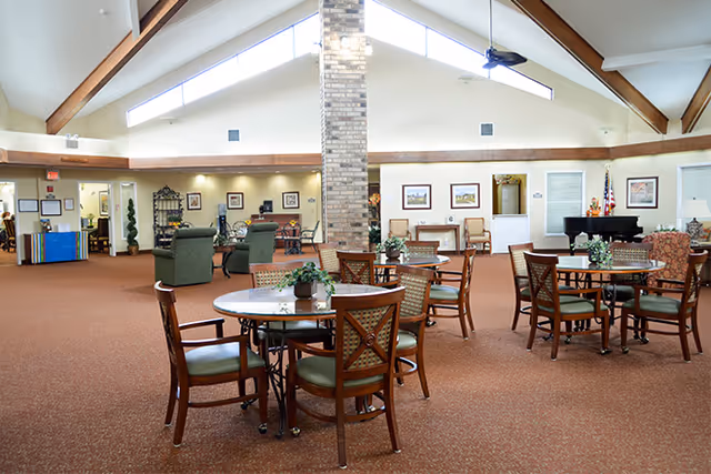 Spacious senior living common room with round tables and chairs, lounge seating, a piano, and a vaulted ceiling with exposed beams.