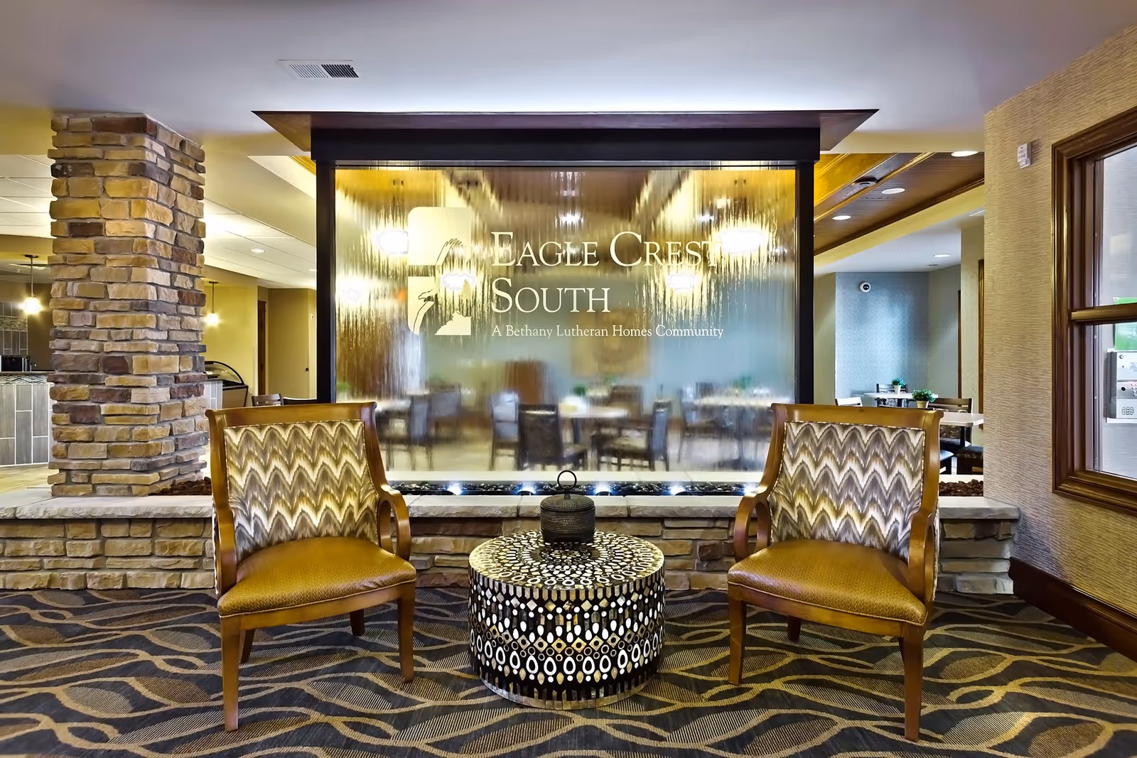 Interior seating area at Eagle Crest South featuring two patterned armchairs and a round decorative table between them, set against a stone wall with a water feature and the Eagle Crest South logo etched on glass behind it. In the background, a dining area with tables and chairs is visible.