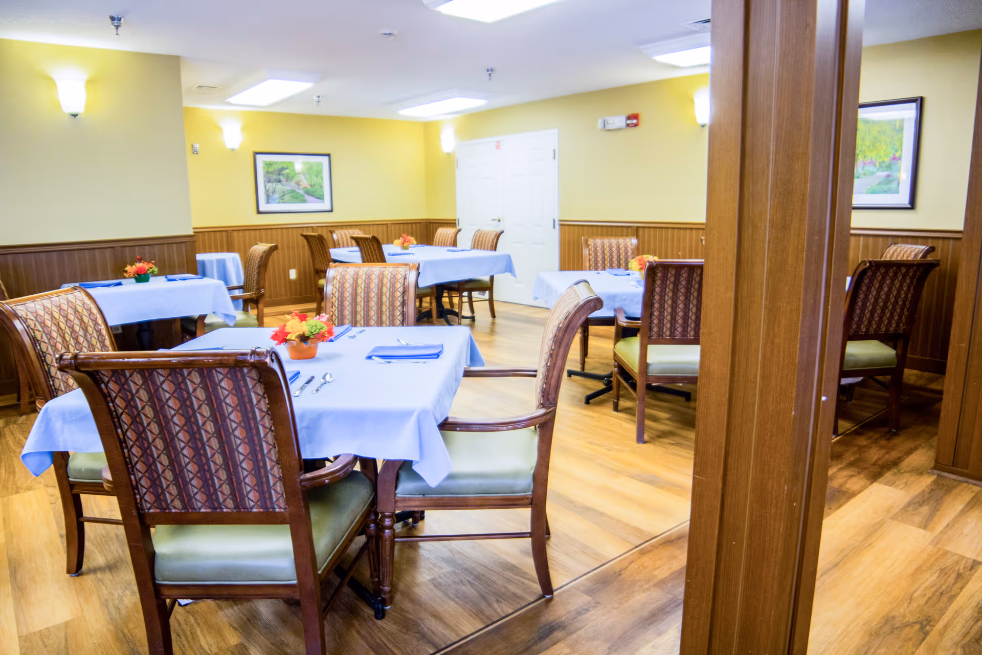 A dining room with multiple tables covered in light blue tablecloths, each set with utensils and small flower arrangements. The room has wooden floors, yellow walls with wood paneling, and framed landscape paintings on the walls. Several upholstered chairs with patterned backs and green seats surround the tables.
