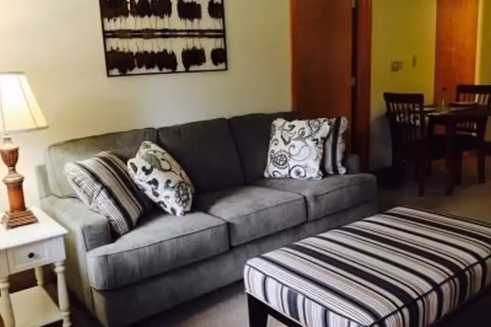 A cozy living room area with a gray sofa adorned with patterned cushions, a striped ottoman in front, a white side table with a lamp, and a dining table with chairs in the background.
