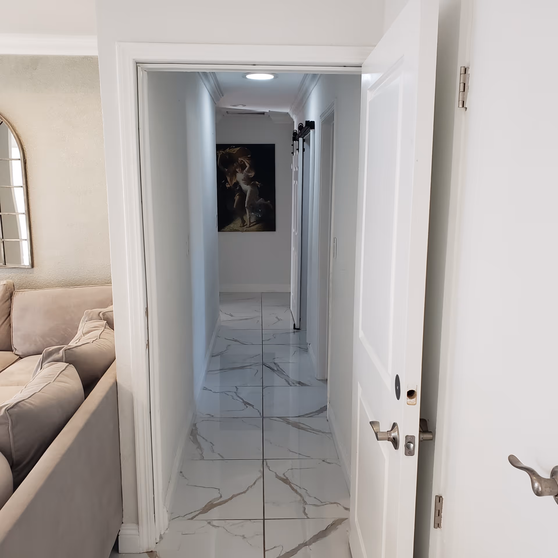 A narrow hallway with white walls and a glossy white marble floor with gray veining. On the left side, there is a beige sectional sofa partially visible with a decorative mirror above it. At the end of the hallway, there is a dark framed artwork on the wall. The hallway has white doors on the right side, one of which is partially open.