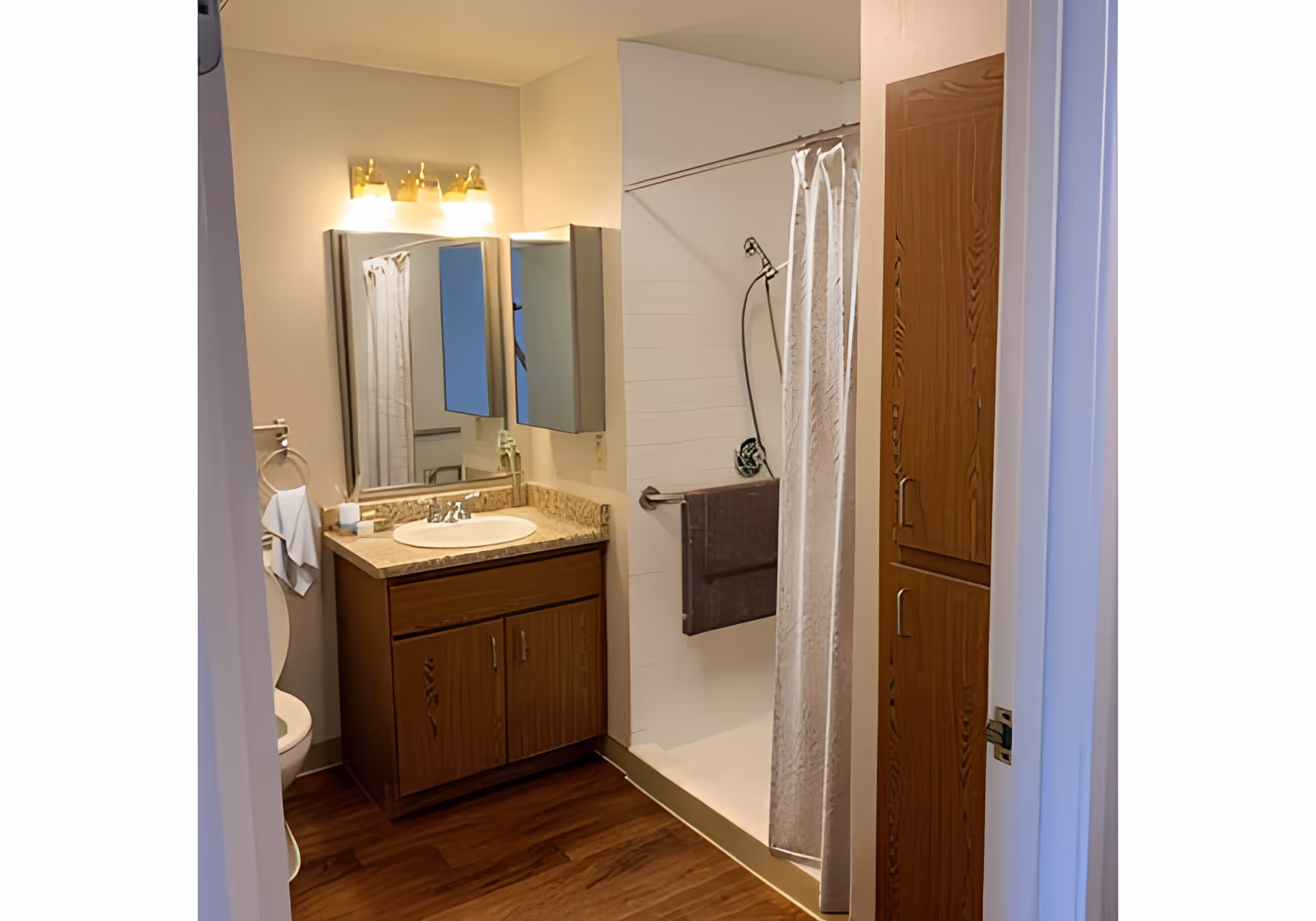 Compact bathroom with a wood vanity and sink under a mirror and light, a shower with a curtain and towel, a toilet partly visible, and tall wooden storage cabinets.