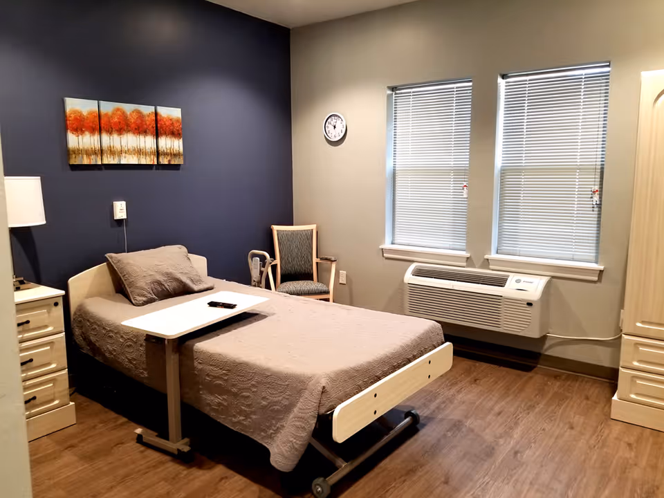 A single bed with a gray quilt and pillow in a room with wooden flooring. There is a small white bedside table with a lamp on the left side of the bed, a chair with a cushion near the corner, and a wall-mounted air conditioning unit below two windows with blinds. A three-panel painting of orange trees hangs on a dark blue accent wall behind the bed.