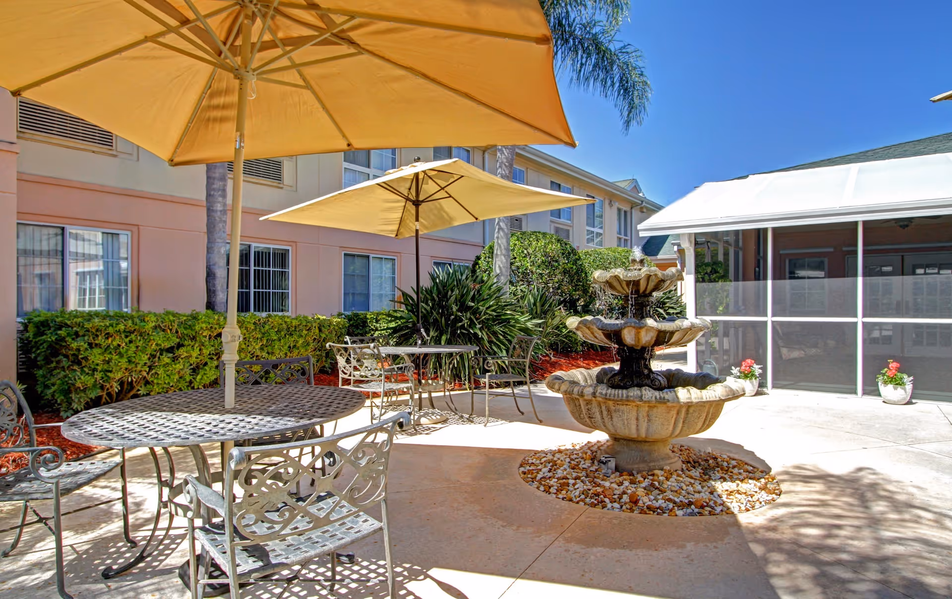 Sunlit outdoor courtyard with metal tables and umbrellas and a tiered fountain in front of a light-colored building.