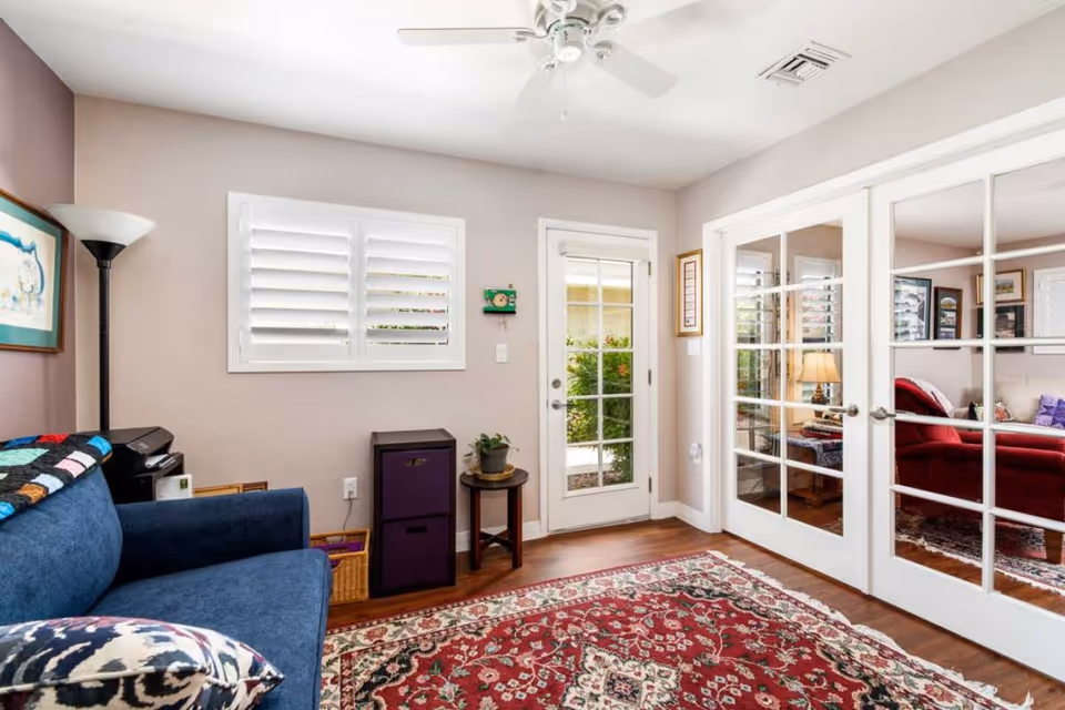 A cozy living room with a blue couch adorned with a colorful quilt and patterned pillow. The room features a red patterned area rug on wooden flooring, a floor lamp, a small table with a plant, and a window with white shutters. There is a glass door leading outside and French doors opening to another room with a red armchair and a lamp.