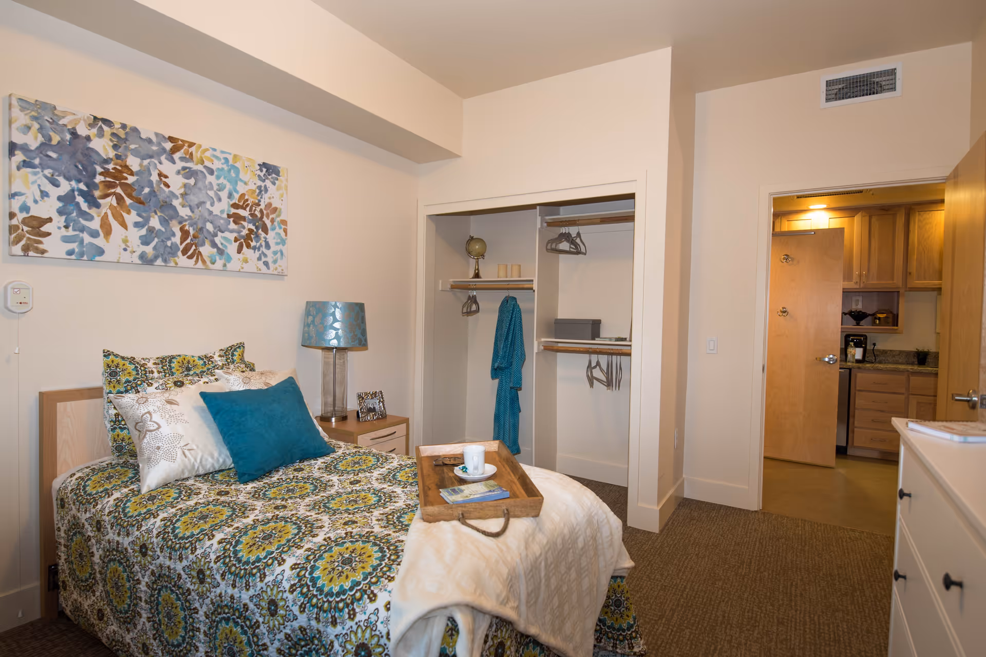 A cozy bedroom with a single bed covered in a patterned green, blue, and yellow bedspread. The bed has several pillows and a wooden tray with a cup and saucer on it. Next to the bed is a nightstand with a blue lamp and a small picture frame. There is an open closet with hangers and a blue robe hanging inside. The room has beige walls and carpeted floor. A doorway leads to a kitchen area with wooden cabinets and a coffee maker visible.