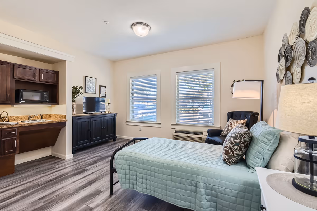 A bright and cozy senior living bedroom featuring a bed with a light green quilt and decorative pillows, a black armchair with a patterned cushion, a floor lamp, and a wall-mounted decorative piece. The room has two windows with blinds, a black cabinet with a TV on top, and a small kitchenette area with a sink, microwave, and dark wood cabinets. The floor is covered with wood-style vinyl flooring.