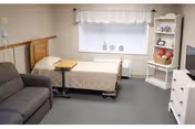 A furnished senior bedroom with a hospital-style bed, gray sofa, window with valance, white shelving and a TV dresser.
