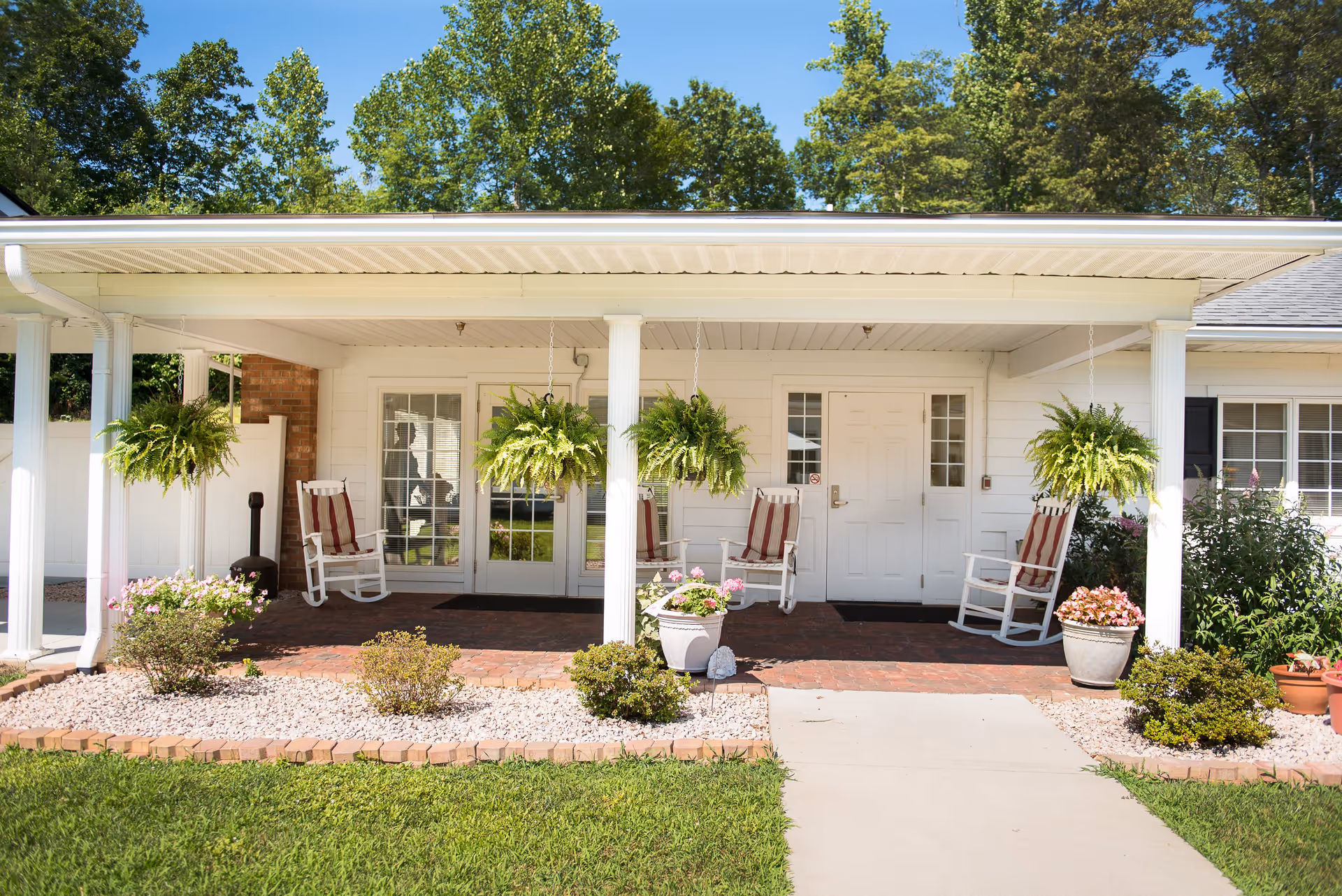 Front porch of a white building with a covered patio featuring four white rocking chairs with red and white striped cushions. Three hanging green ferns are suspended from the ceiling. There are potted plants and small bushes arranged in a landscaped area with white rocks and brick edging in front of the porch. The building has white double doors and windows, and there are trees visible in the background under a clear blue sky.