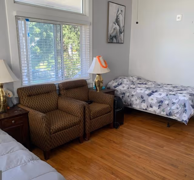 A bright room with two patterned armchairs by a large window, lamps and a single bed with floral bedding.