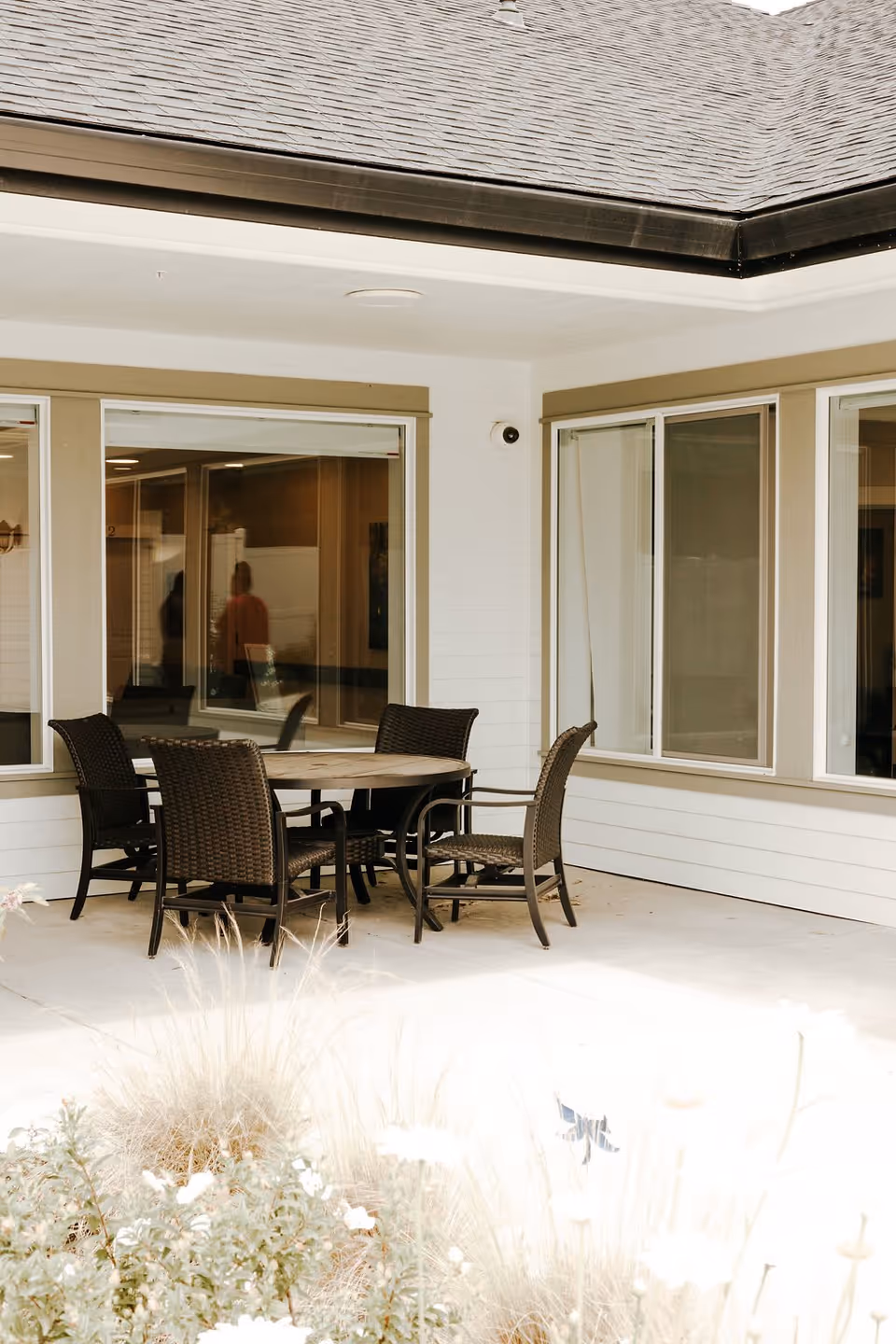 Outdoor patio area at Kinsington Oak Grove with a round table and four wicker chairs on a concrete floor, surrounded by windows and beige siding, with some plants and flowers in the foreground.
