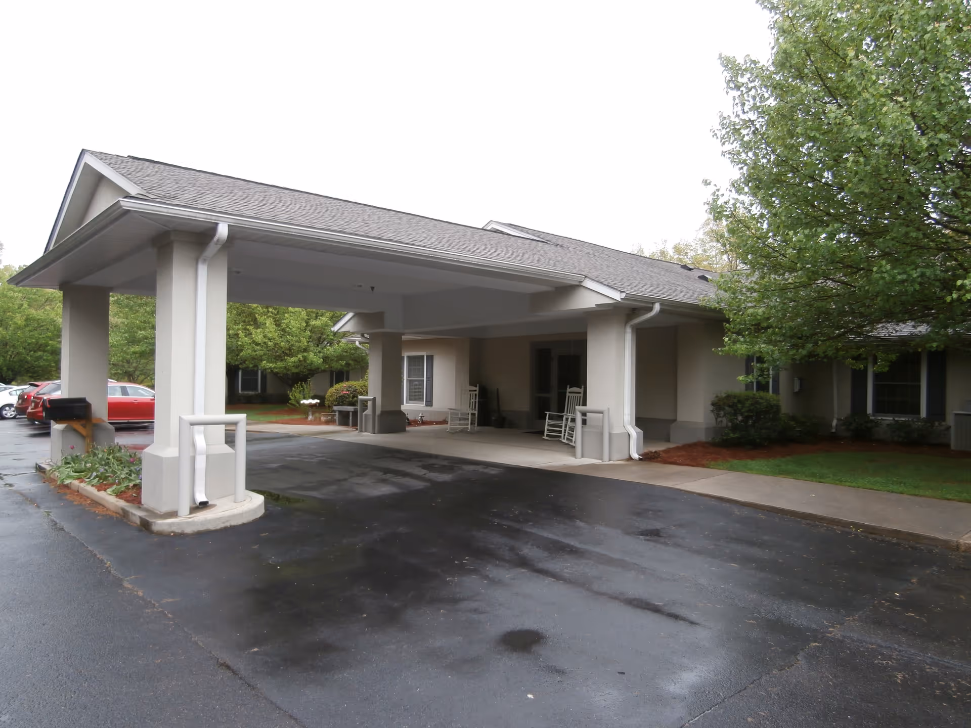 Covered porte-cochere and entrance of a single-story senior living building with rocking chairs and wet pavement.