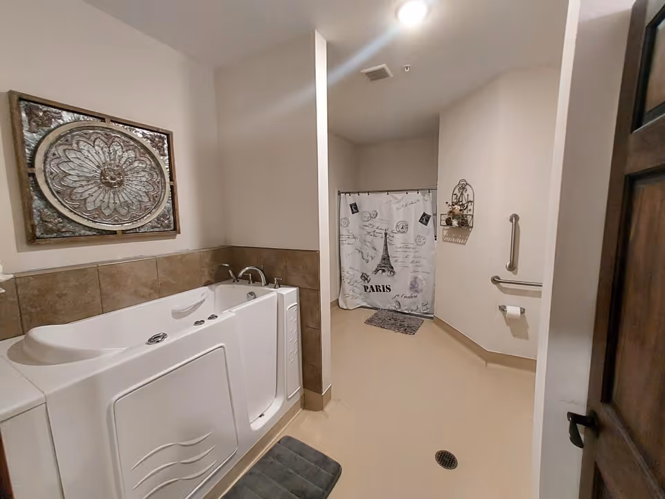 A spacious bathroom featuring a walk-in bathtub with a tiled surround on the left and a shower area with a Paris-themed curtain in the back. The walls are light-colored, and there is a decorative wall hanging above the bathtub. Safety grab bars and a toilet paper holder are mounted on the right wall. The floor is smooth and light-colored with a drain in the center.