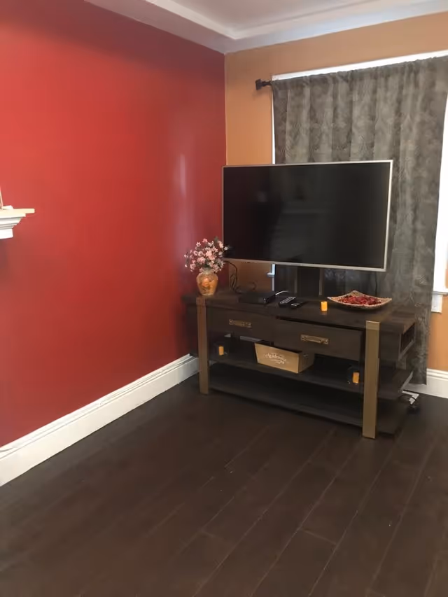 A corner of a living room with a red and orange painted wall, a dark wooden TV stand holding a flat-screen television, a vase with flowers, remote controls, and decorative items. There is a window with patterned curtains behind the TV stand and dark wooden flooring.
