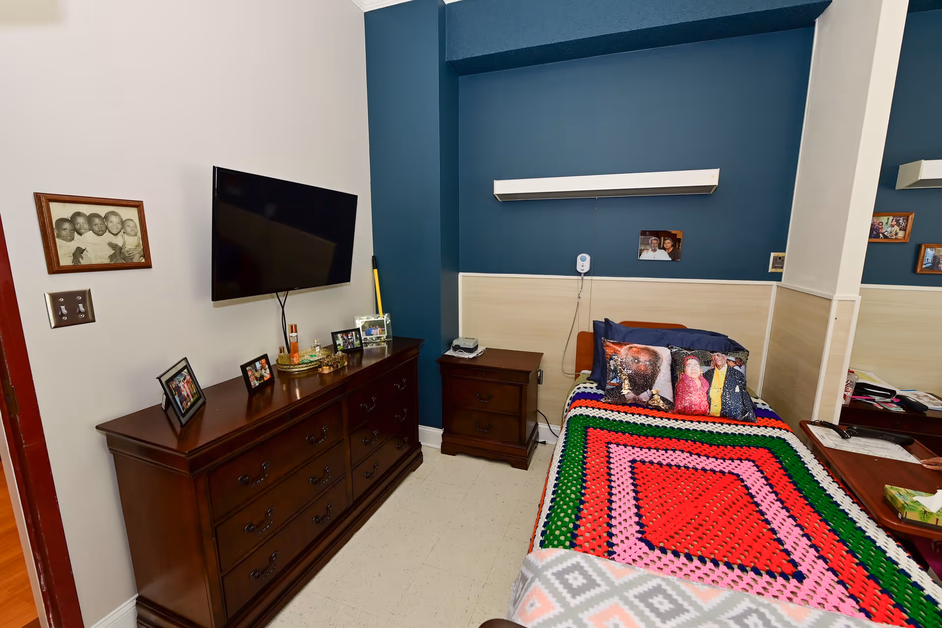 A small bedroom with a single bed covered in a colorful crocheted blanket and pillows featuring printed photos. There is a dark wooden dresser with framed photos and a flat-screen TV mounted on the wall above it. A nightstand with a phone and other items is next to the bed. The walls are painted white and dark blue, with some family photos hanging on the walls.