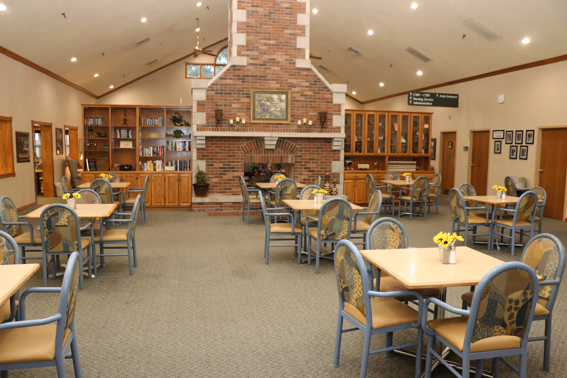 A spacious dining room with multiple square tables and chairs arranged neatly. Each table has a small vase with yellow flowers. The room features a large brick fireplace in the center of the far wall, flanked by wooden bookshelves and cabinets. The ceiling is vaulted with recessed lighting, and there are framed pictures on the walls.