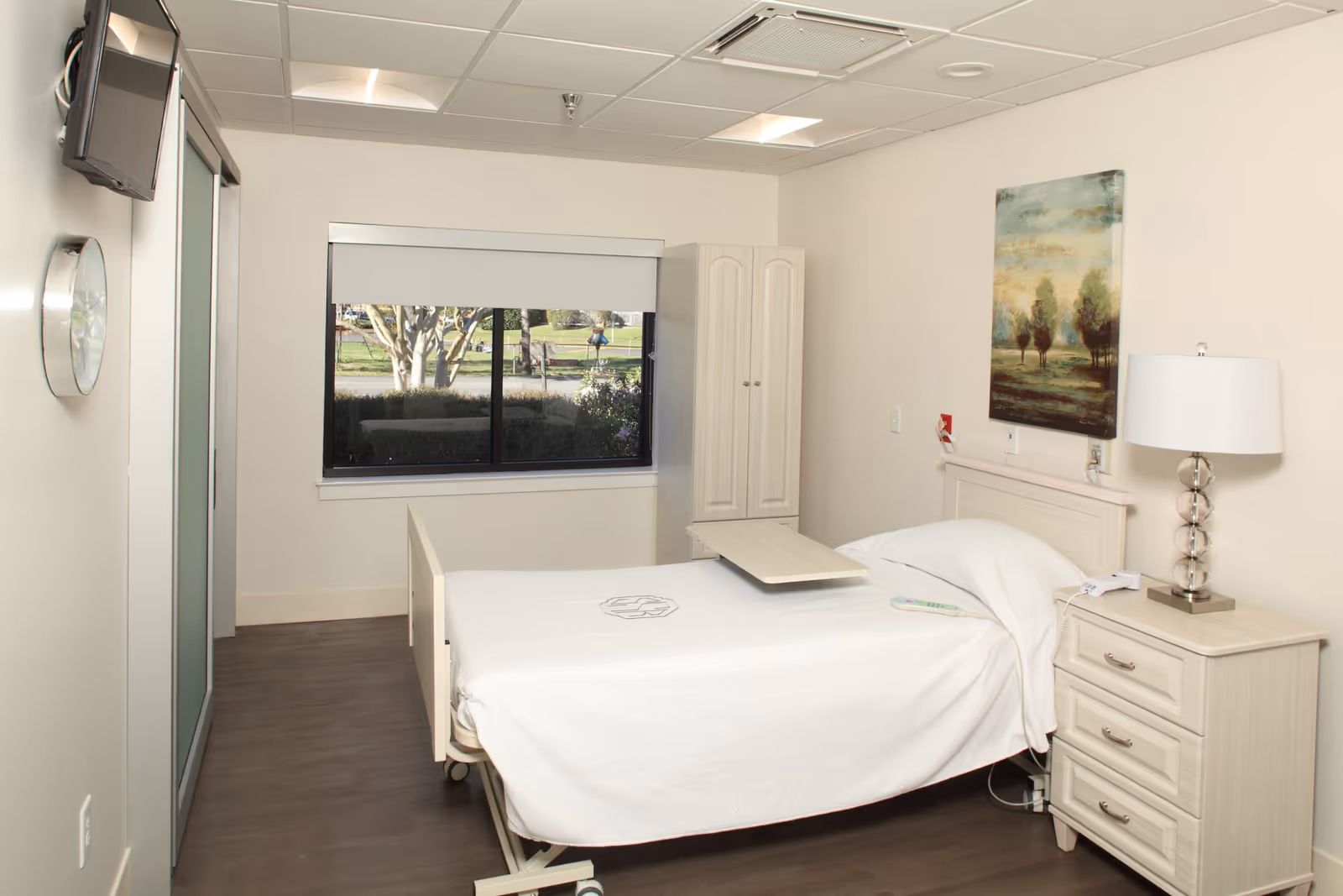 A clean and bright senior residence bedroom with a single hospital-style bed covered in white linens. The room features a bedside table with a lamp, a small cabinet, a wall-mounted TV, a clock, and a window with a view of greenery outside. There is a piece of artwork hanging above the bed.