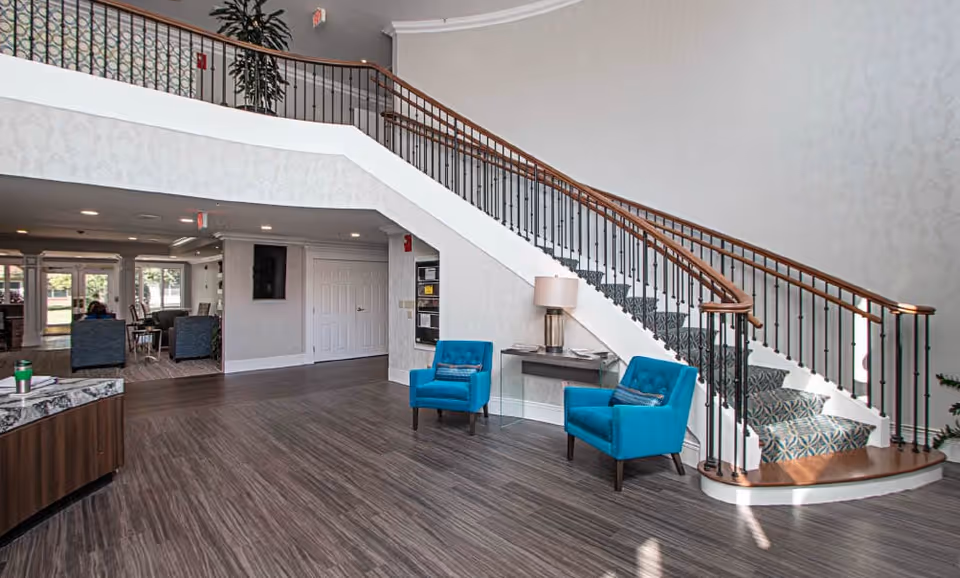 Spacious interior lobby with a sweeping curved staircase, two bright blue chairs, a small table with a lamp, and wood-tone flooring.
