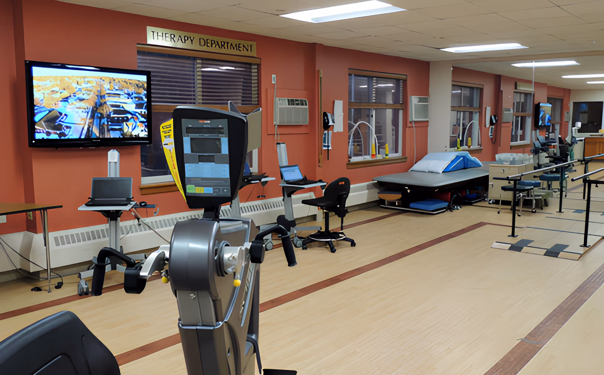 Interior view of a therapy department room in a rehabilitation and nursing center, featuring exercise equipment, therapy tables, chairs, wall-mounted televisions, and large windows with blinds. The room has a wooden floor with marked lines and a large mirror on one wall.