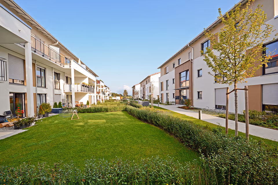 Courtyard with a green lawn and hedges between modern multi-story senior living apartment buildings under a clear blue sky.