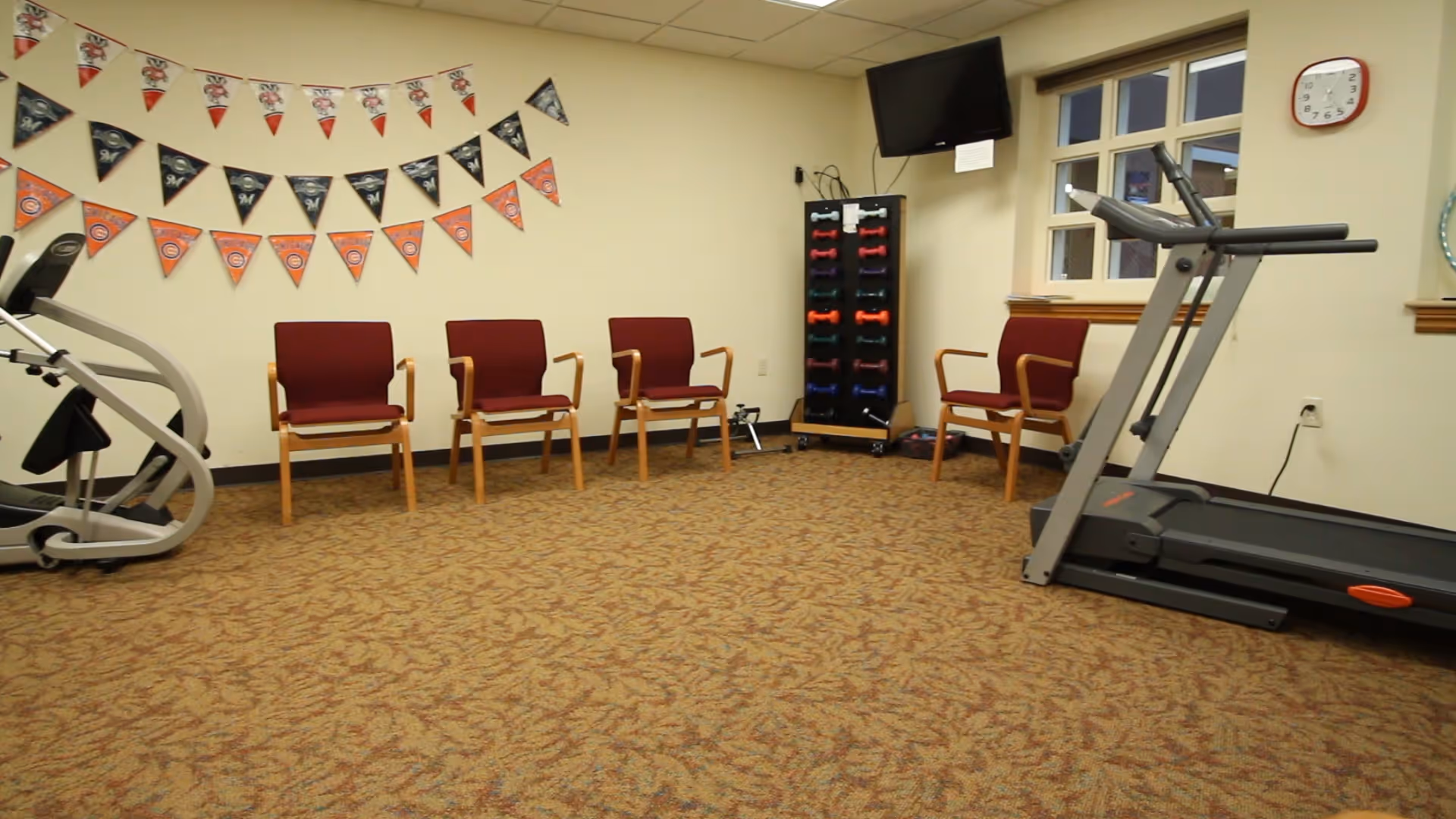 A small exercise room with a treadmill, an elliptical machine, four maroon chairs with wooden arms, a rack of colorful dumbbells, a wall-mounted TV, a clock, and decorative pennant banners on the wall.