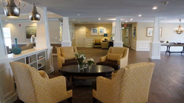 Bright communal lounge with four patterned armchairs around a round coffee table in a spacious senior living common area.