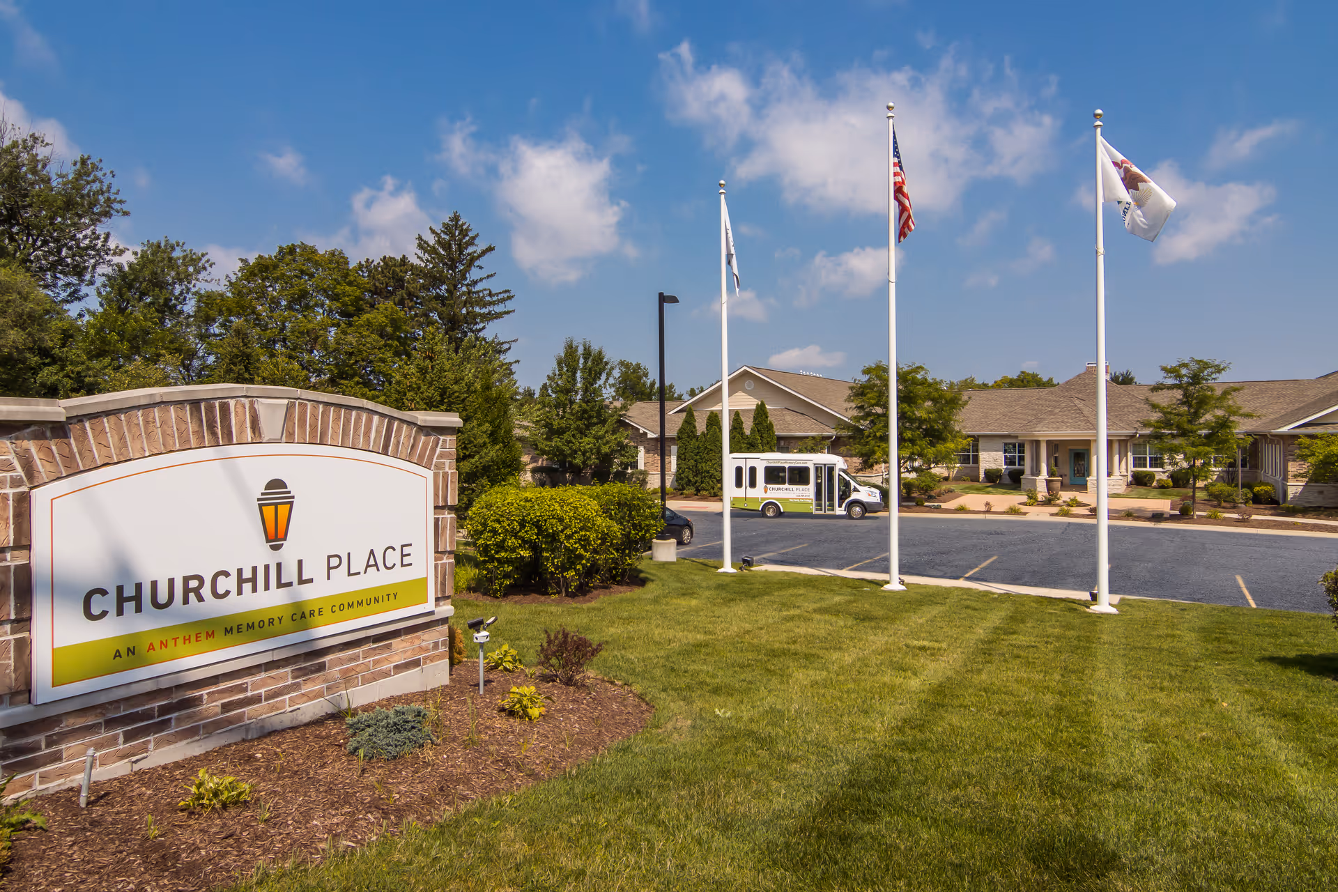 Exterior view of Churchill Place Memory Care facility with a large sign in the foreground, three flagpoles with flags, a parking lot, and a building entrance under a blue sky with some clouds.