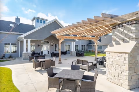 Outdoor patio area at Villas of Holly Brook Assisted Living & Memory Care in Bellevue, IL, featuring multiple wicker tables and chairs arranged on a concrete surface. A wooden pergola provides partial shade, and a stone fireplace is visible on the right. The building exterior is gray with white trim and several windows, under a blue sky with some clouds.