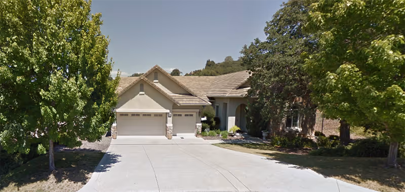 Front exterior view of a single-story residential building with a three-car garage, surrounded by trees and greenery under a clear sky.