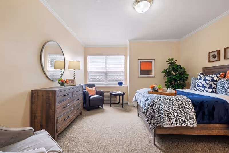 A well-lit bedroom with beige walls and carpeted floor featuring a wooden bed with blue and white patterned bedding and multiple pillows. A wooden dresser with a round mirror and a table lamp is on the left side. There is a dark armchair with an orange pillow and a small round table near a window with blinds. A green potted plant and framed artwork decorate the room.
