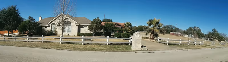Wide panoramic view of a single-story residential building with a light-colored exterior, surrounded by a white wooden fence and a mailbox in the foreground. The property has a driveway, some trees, and a clear blue sky above.