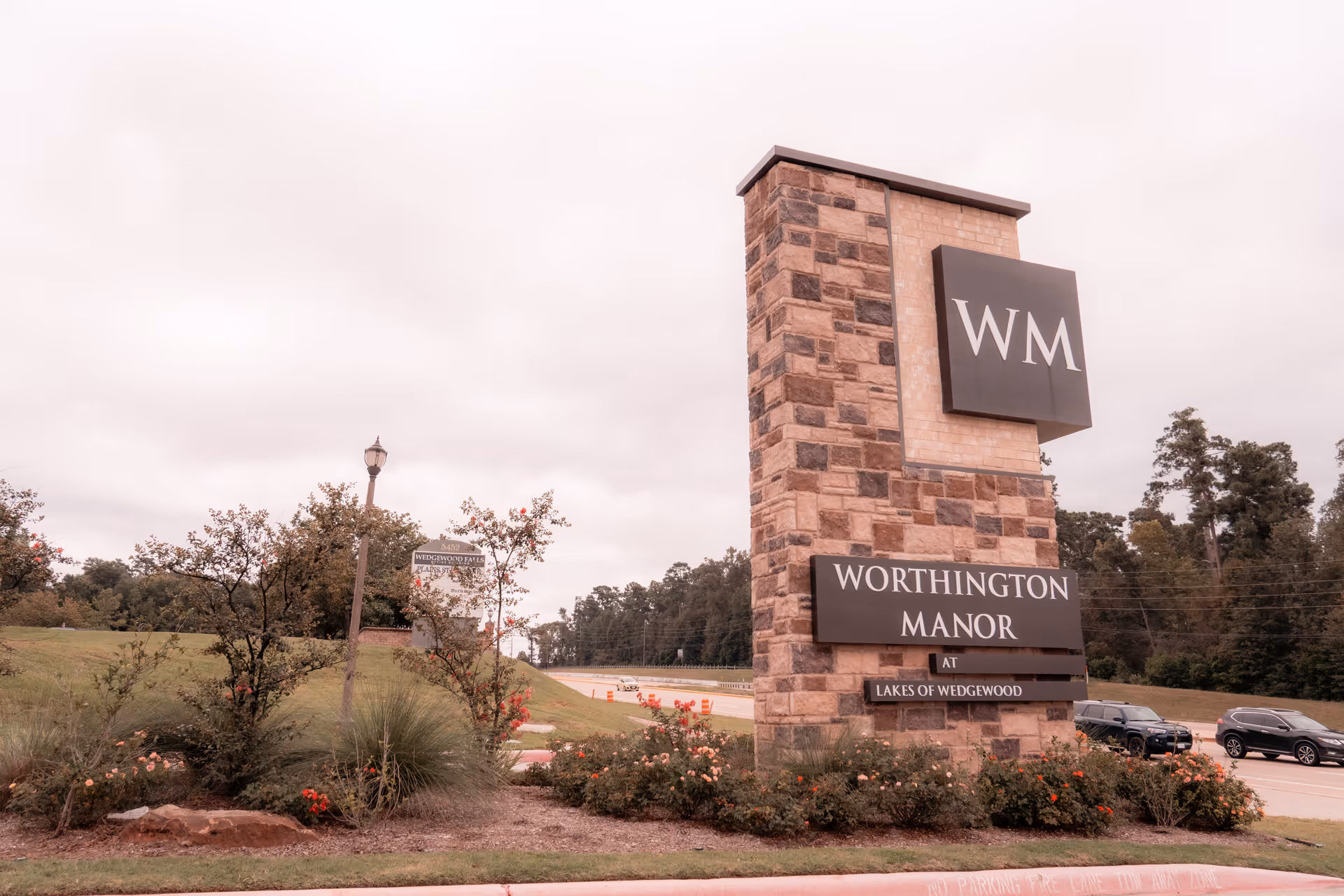 A large stone sign for Worthington Manor at Lakes of Wedgewood stands beside a road with cars parked nearby. The sign features the initials WM at the top and is surrounded by landscaping with bushes and flowers. Trees and a cloudy sky are visible in the background.